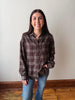 A woman with long dark hair stands indoors wearing a brown plaid button-up shirt and blue jeans, with a plain off-white wall and wooden floor behind her.