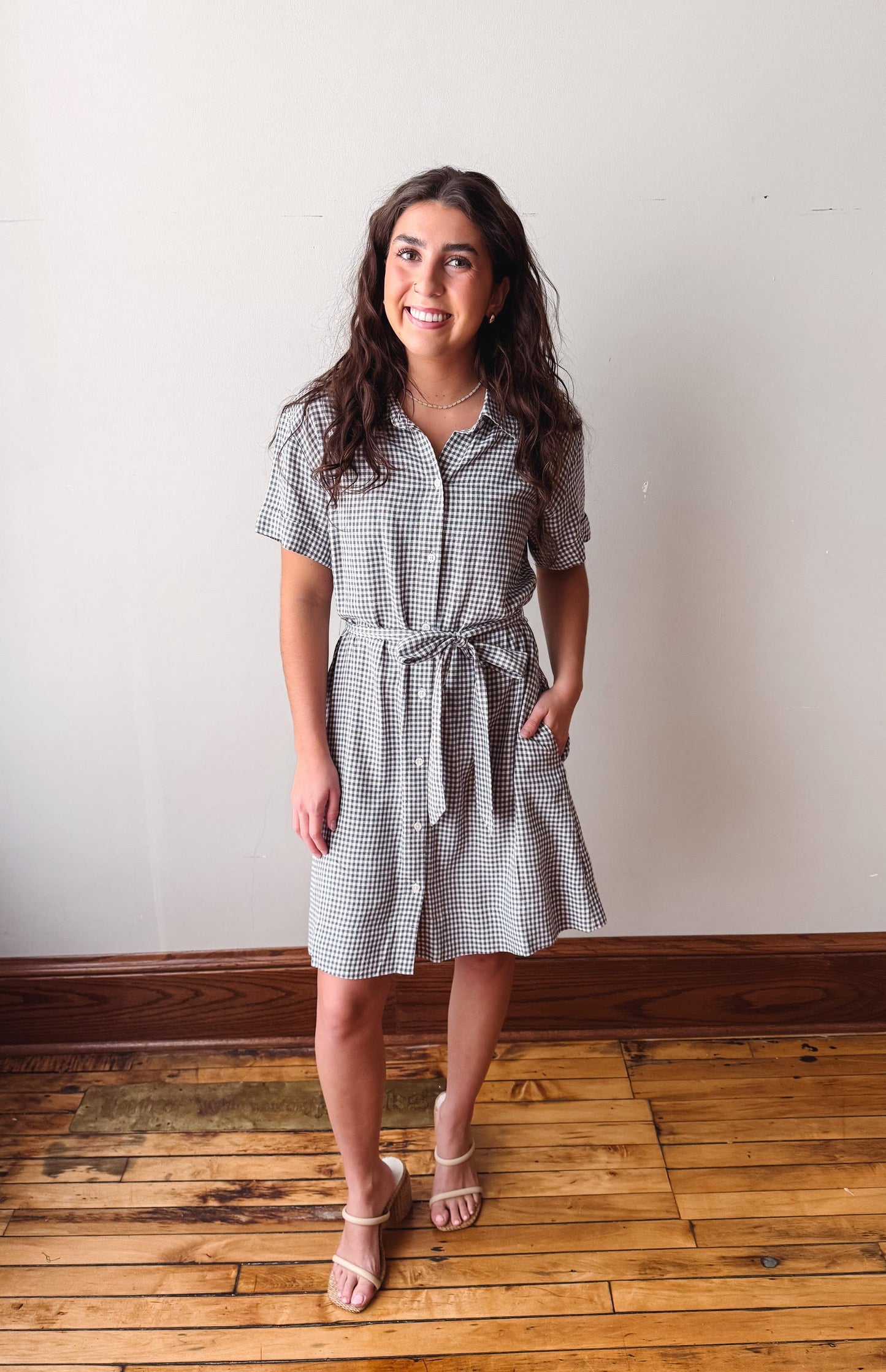 Woman wearing a checkered dress standing on a wooden floor with a white wall background