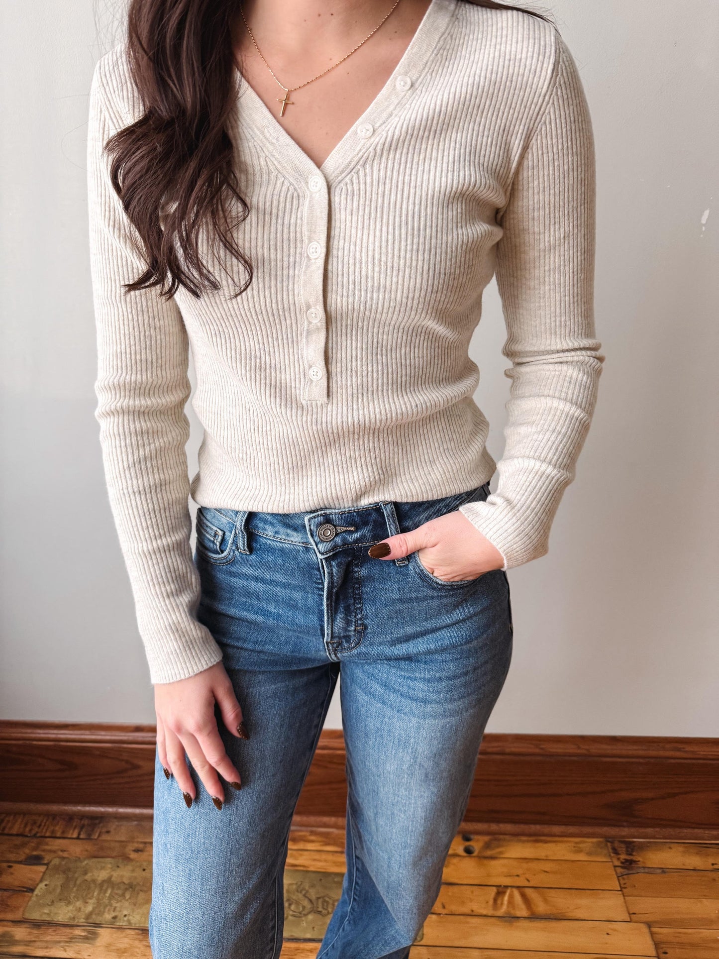 A woman wearing a cream ribbed long-sleeve button-front cardigan sweater and blue jeans, standing indoors against a light wall with a wood floor visible.