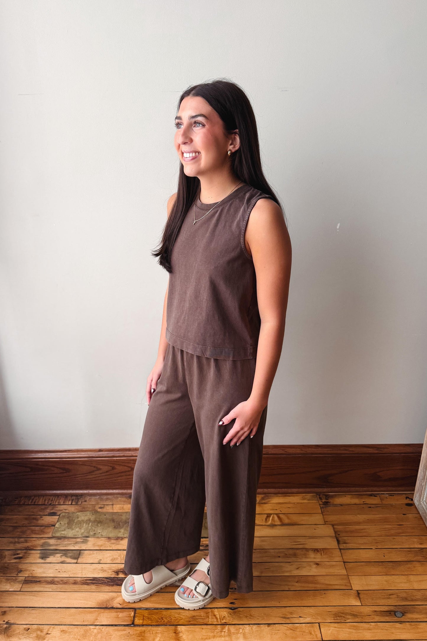 A woman modeling a sleeveless brown jumpsuit with wide-leg pants, standing on a wooden floor against a plain light gray wall.