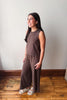 A woman modeling a sleeveless brown jumpsuit with wide-leg pants, standing on a wooden floor against a plain light gray wall.