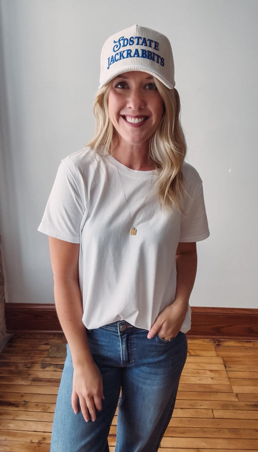 A smiling blonde woman wearing a white t-shirt, blue jeans, and a white baseball cap with blue 'NSSTATE JACKRABITS' lettering, standing in a room with a light gray wall and wooden floor.