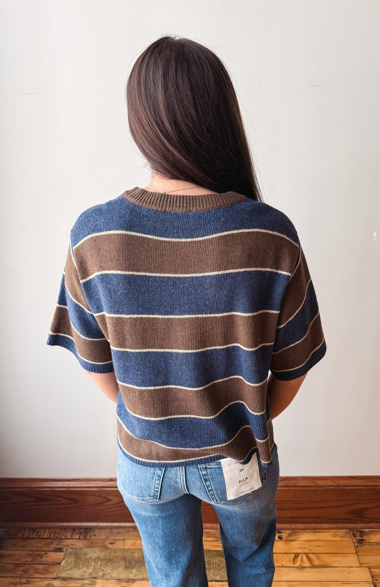 A woman viewed from behind wearing a short-sleeve knit sweater with horizontal stripes in navy blue, brown, and cream, paired with blue jeans, standing against a plain off-white wall.