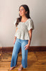 Woman wearing a light green blouse with ruffled sleeves and blue jeans standing on a wooden floor.