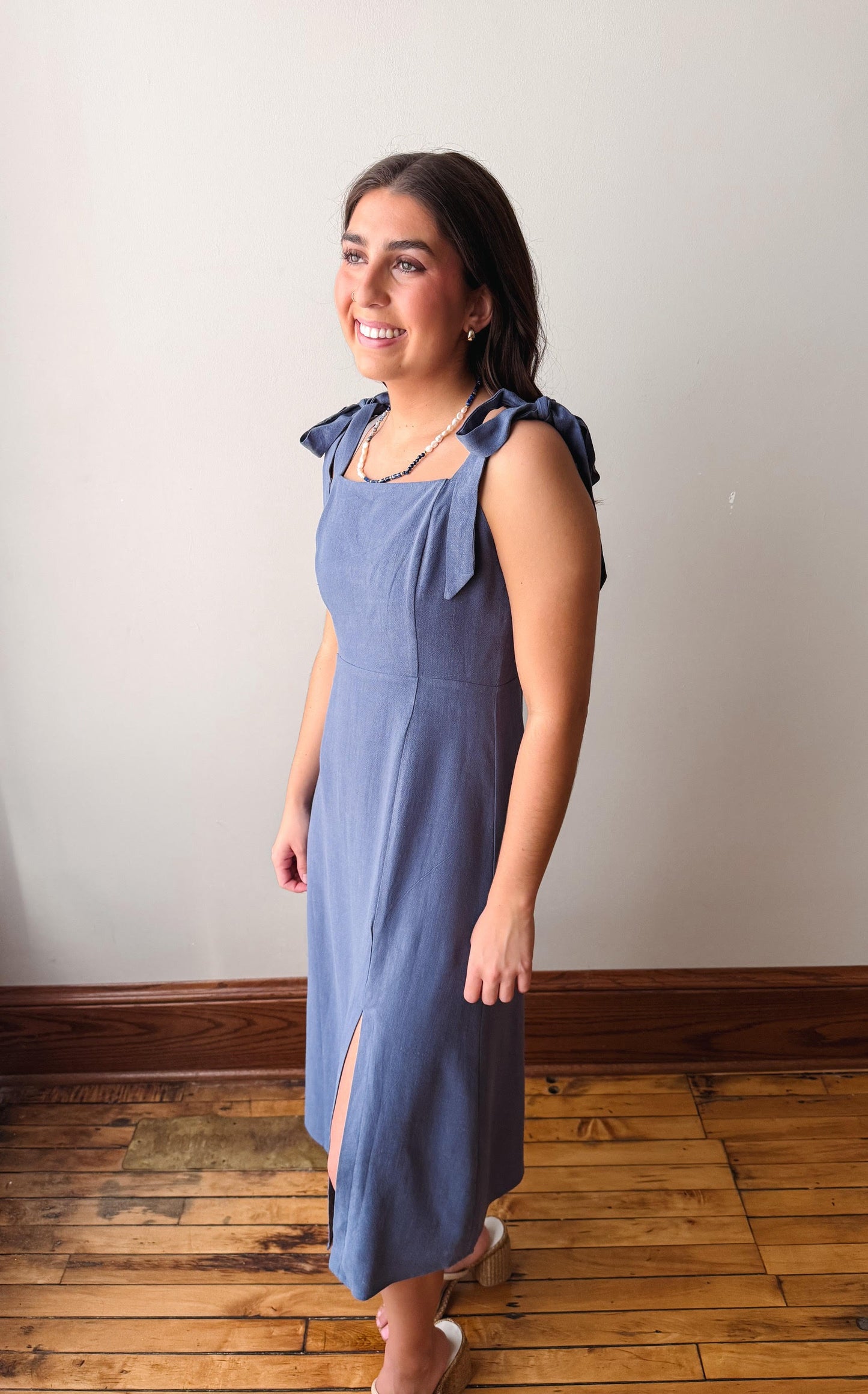 Woman wearing a blue dress with bow straps standing on a wooden floor against a white wall.