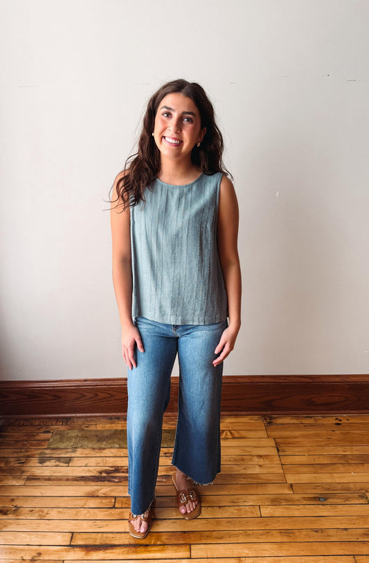 Woman wearing a light blue sleeveless top and jeans standing on a wooden floor.