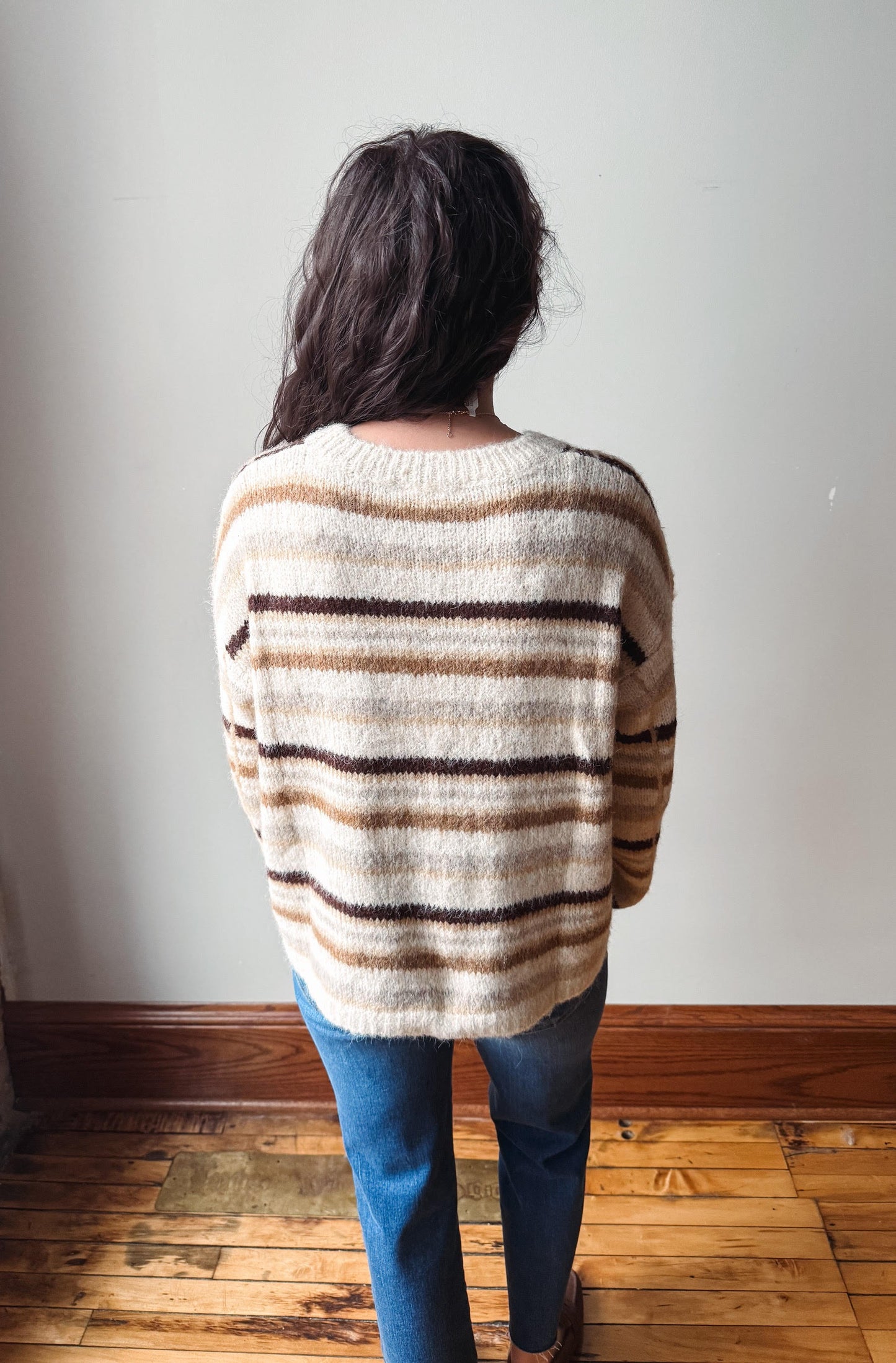 A person viewed from behind wearing a fuzzy knit sweater with horizontal beige, brown, and dark brown stripes, paired with blue jeans, standing on a wooden floor against a plain wall.