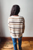 A person viewed from behind wearing a fuzzy knit sweater with horizontal beige, brown, and dark brown stripes, paired with blue jeans, standing on a wooden floor against a plain wall.
