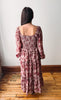 A woman viewed from behind wearing a dusty rose and burgundy floral tiered maxi dress with long puff sleeves and a smocked bodice, standing on a wooden floor against a plain white wall.