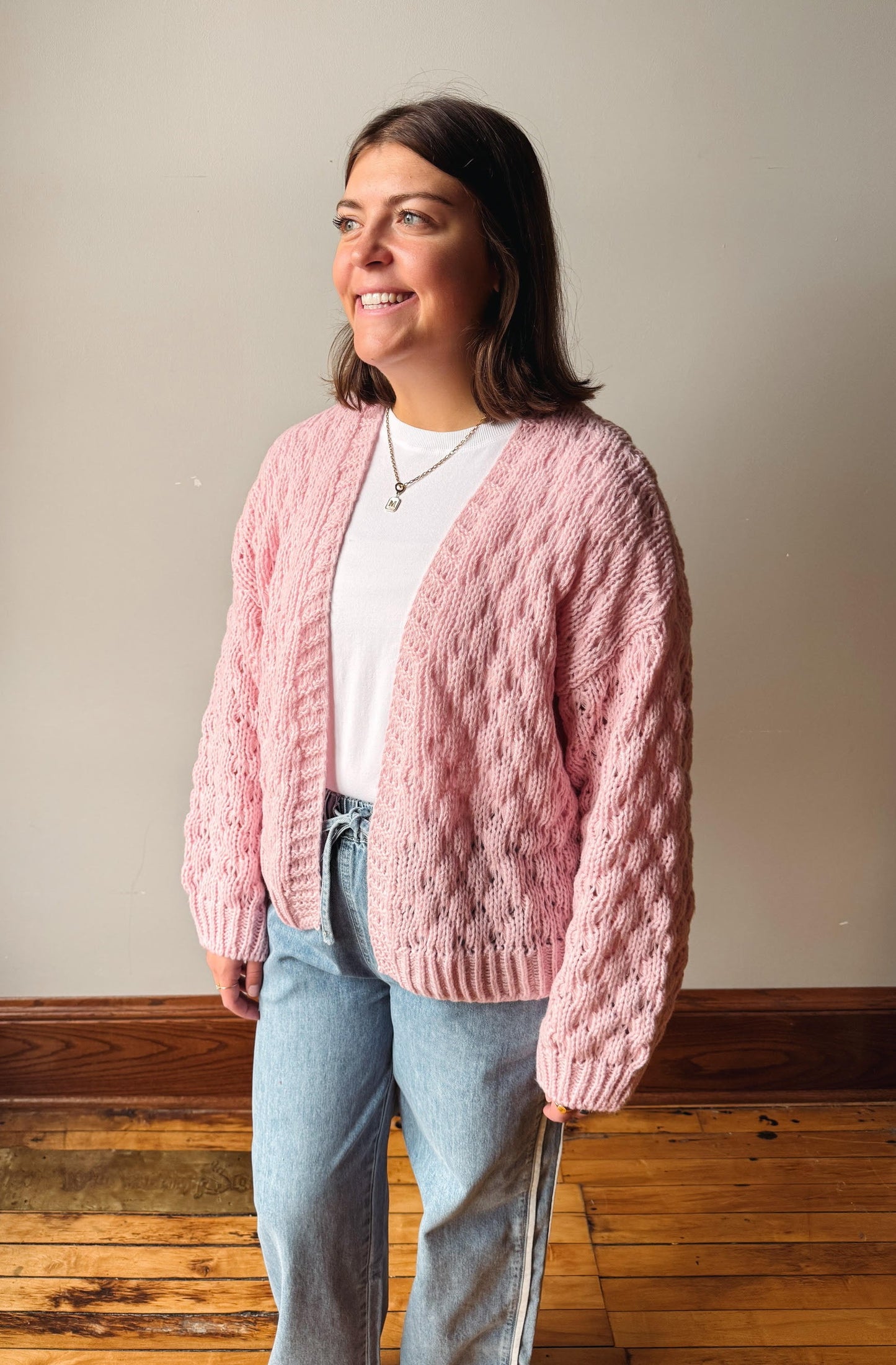 A woman wearing a chunky pink cable-knit cardigan over a white t-shirt and light blue jeans, standing in a room with a beige wall and wooden floor.