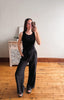Woman taking a mirror selfie wearing a black sleeveless top and dark pants in a room with wooden flooring.
