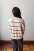 A person viewed from behind wearing a cream-colored knit sweater with thin black horizontal stripes and dark gray pants, standing in an indoor setting with a light gray wall and wooden floor.
