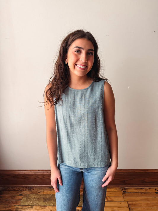 Woman wearing a light blue sleeveless top and jeans standing against a plain wall.