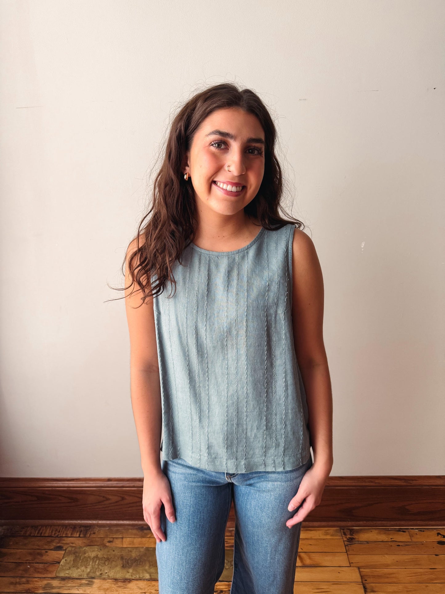 Woman wearing a light blue sleeveless top and jeans standing against a plain wall.