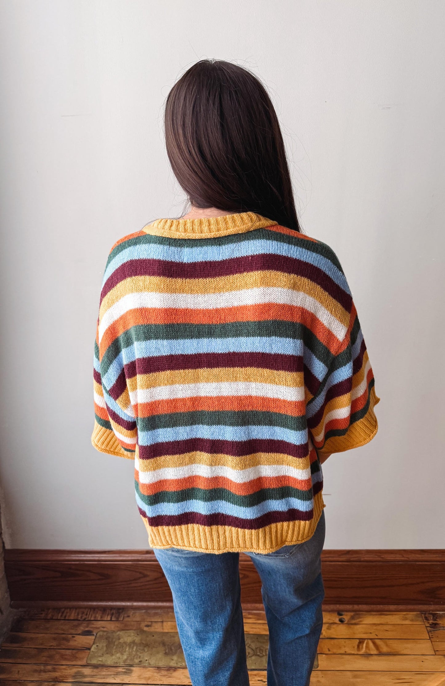 A person viewed from behind wearing a loose, short-sleeve knit sweater with horizontal stripes in yellow, orange, burgundy, green, light blue, and white, paired with blue jeans against an indoor wall and wood floor.