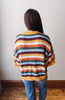 A person viewed from behind wearing a loose, short-sleeve knit sweater with horizontal stripes in yellow, orange, burgundy, green, light blue, and white, paired with blue jeans against an indoor wall and wood floor.