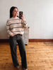 A woman takes a mirror selfie wearing a beige mock-neck sweater with black horizontal stripes, dark charcoal wide-leg pants, and brown boots, standing in a room with a light gray wall and wooden floor.