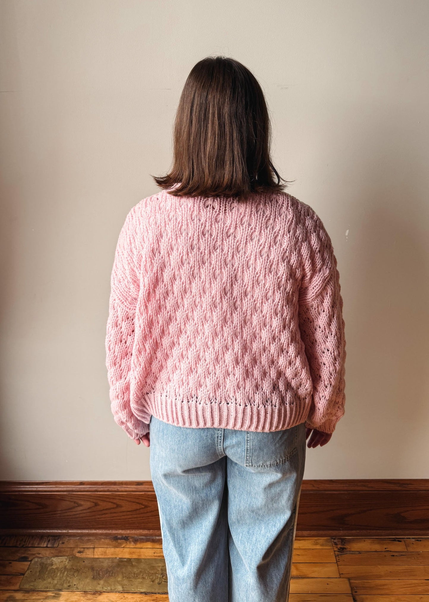 A person viewed from behind wearing a pink chunky cable-knit sweater and light blue jeans, standing in front of a plain beige wall with a wooden baseboard and floor.