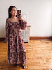 A woman takes a mirror selfie wearing a long-sleeved, tiered maxi dress with a dusty rose and burgundy floral pattern on a beige background, standing on a wooden floor in front of a white wall.