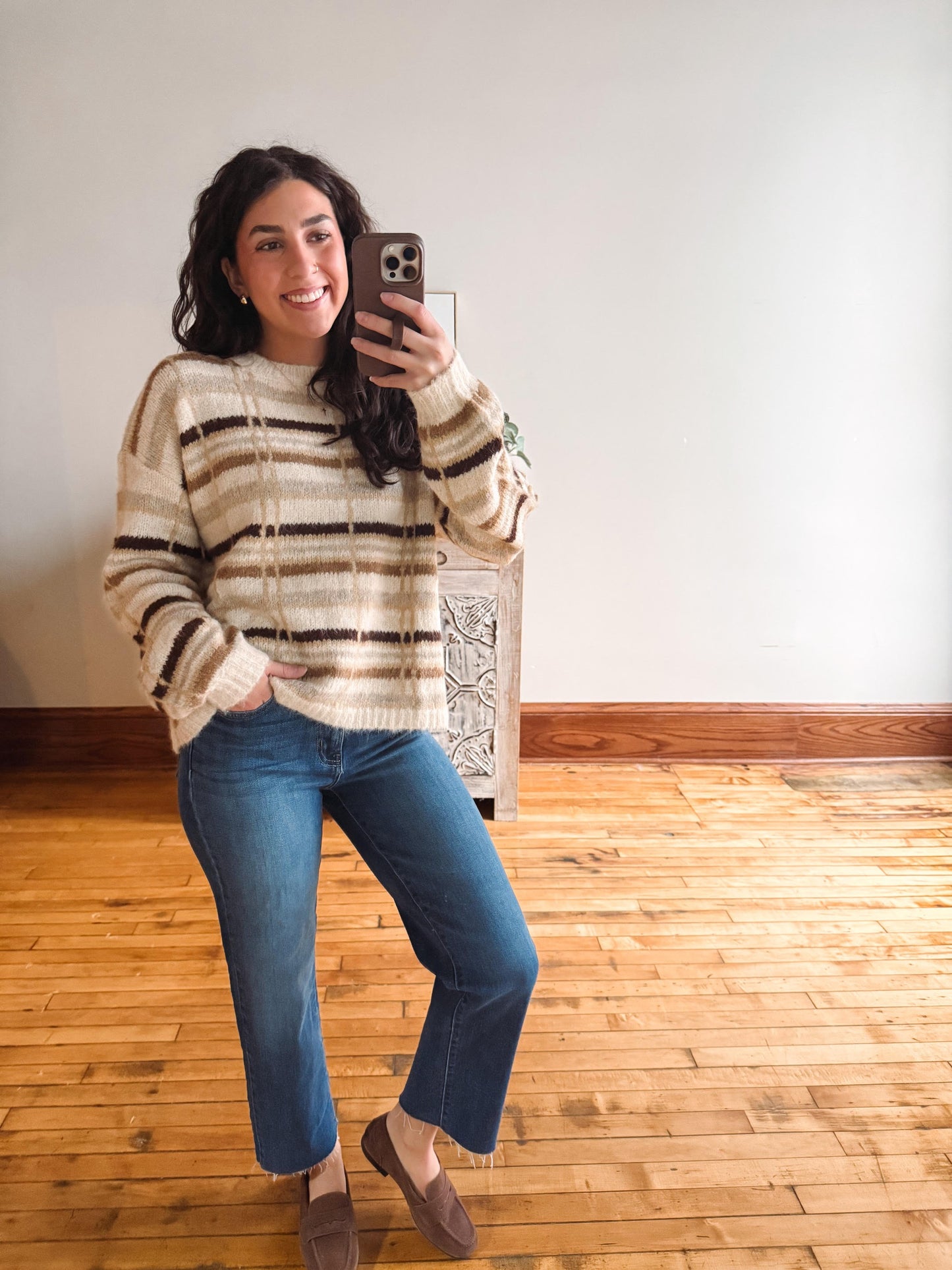 A woman takes a mirror selfie wearing a cream and brown striped knit sweater, blue jeans, and brown slip-on shoes in a room with a white wall and wooden floor.