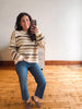 A woman takes a mirror selfie wearing a cream and brown striped knit sweater, blue jeans, and brown slip-on shoes in a room with a white wall and wooden floor.
