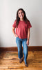 A woman stands in a room with a white wall and wooden floor, wearing a muted red short-sleeve t-shirt, blue jeans, and brown shoes.