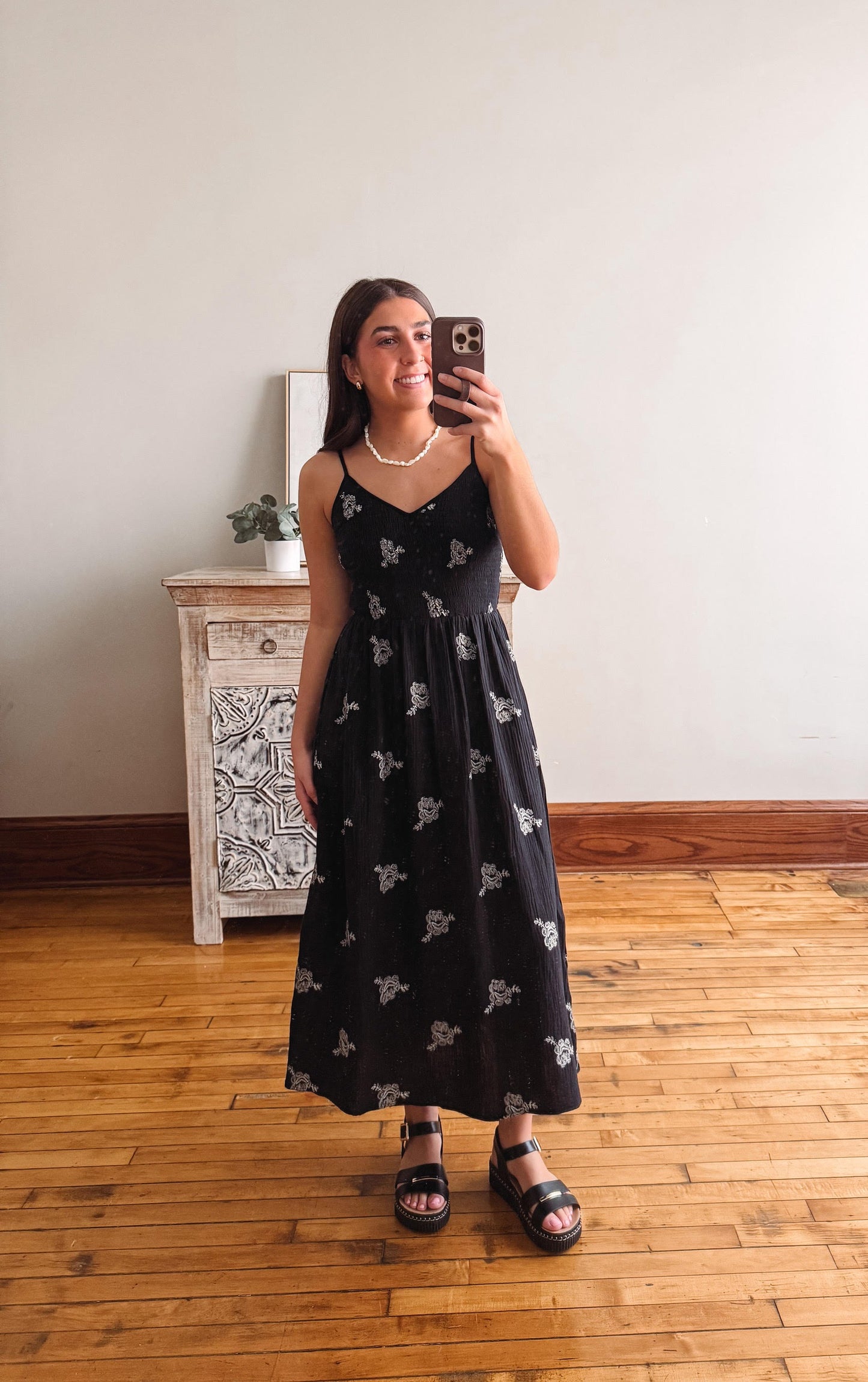 Woman taking a mirror selfie wearing a black dress with white patterns in a room with wooden flooring and a white wall.