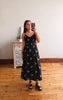 Woman taking a mirror selfie wearing a black dress with white patterns in a room with wooden flooring and a white wall.