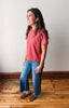 A woman modeling a short-sleeve crewneck t-shirt in a dusty coral pink color, paired with blue jeans and brown suede loafers, standing on a wooden floor against a plain off-white wall.