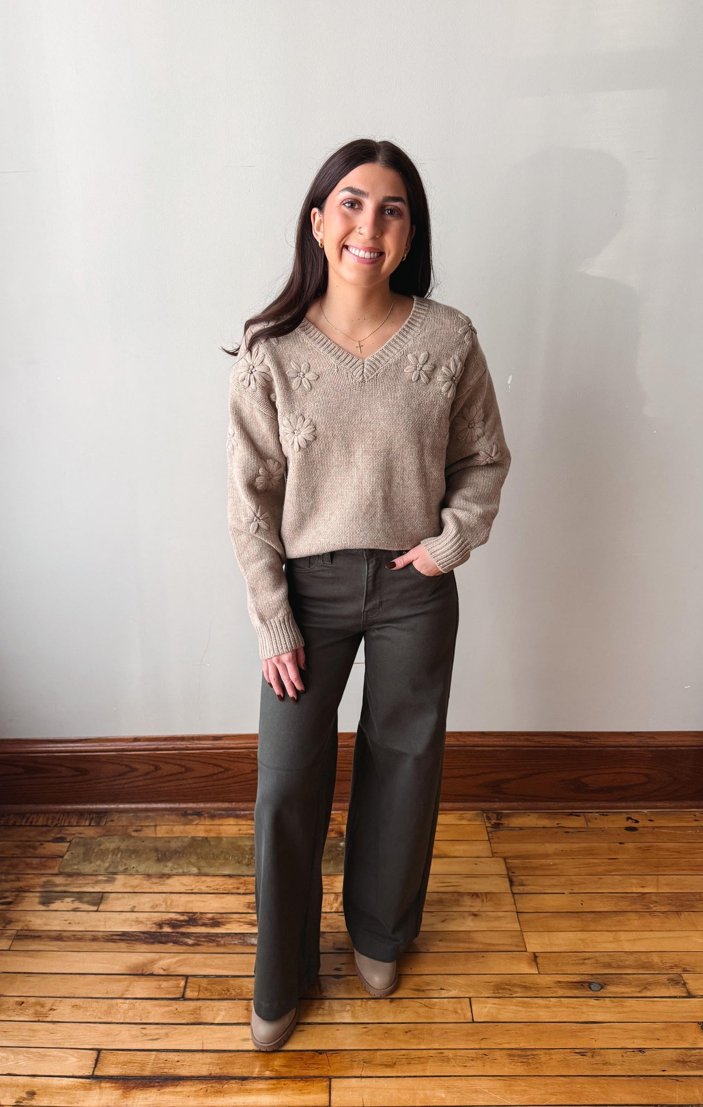 A woman stands in a room with a light gray wall and wooden floor, wearing a beige knit sweater with floral embroidery and dark charcoal wide-leg pants.