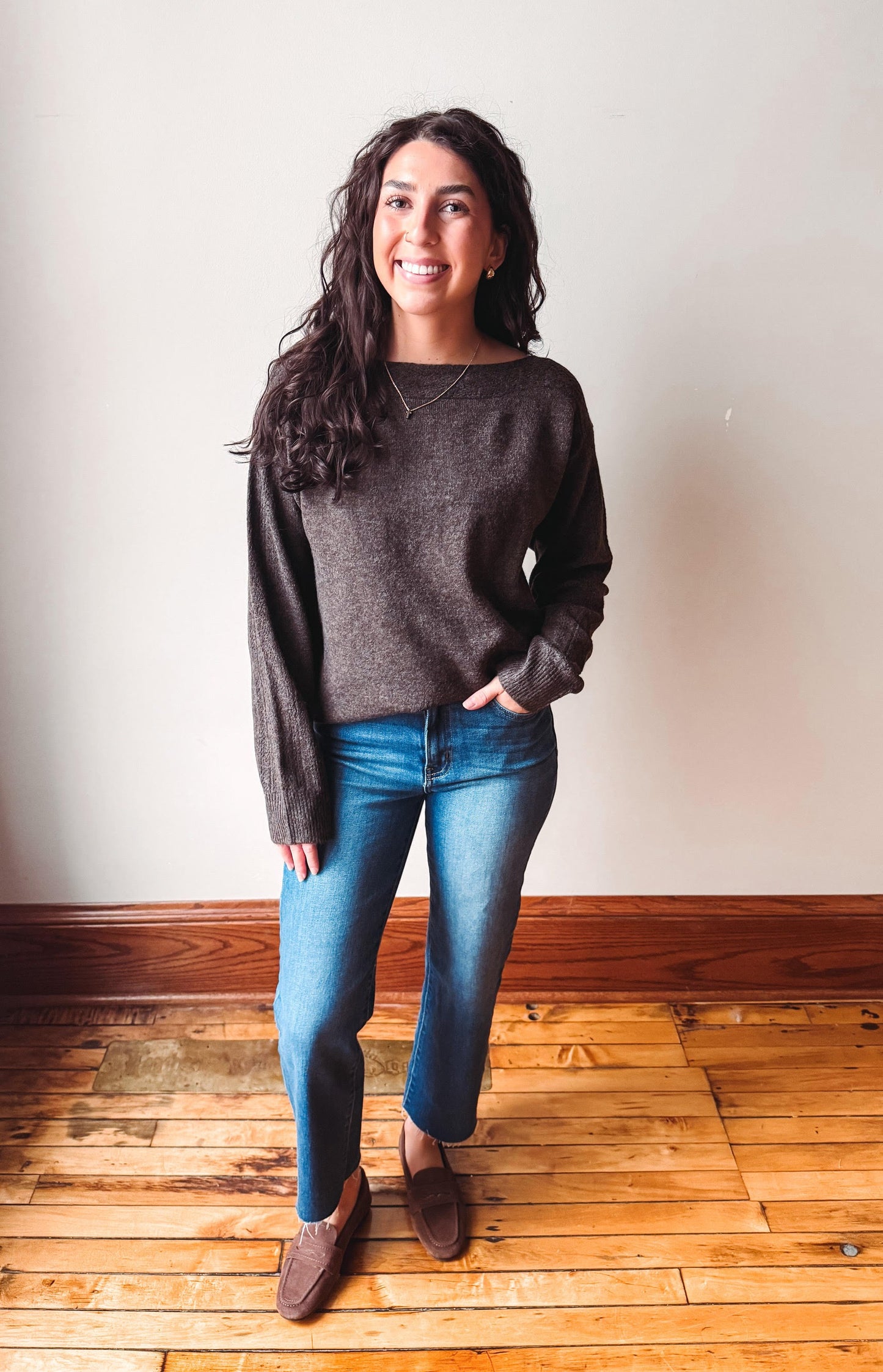 A woman stands in a room with a light beige wall and wooden floor, wearing a dark brown knit sweater, blue jeans, and brown leather loafers.