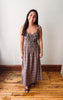 A woman modeling a brown and beige plaid maxi dress with a smocked bodice, standing on a wooden floor against a plain off-white wall.