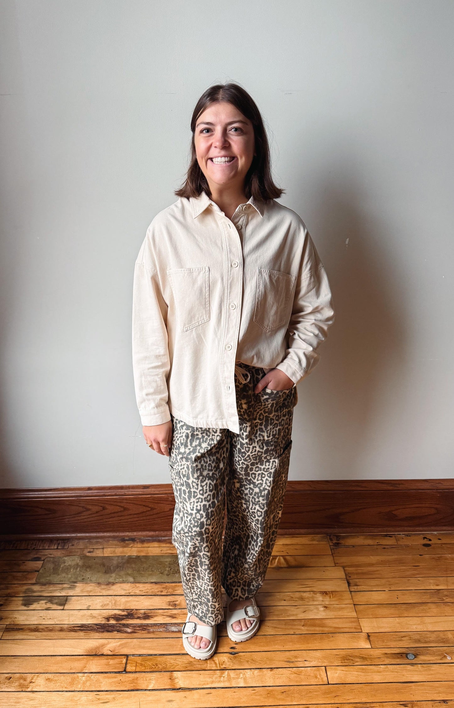 A woman stands in a room with a light gray wall and wooden floor, wearing a cream-colored long-sleeve button-down shirt and leopard-print pants.