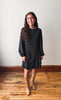 A woman stands in a room with a light beige wall and wooden floor, wearing a black long-sleeve mini dress with a small white pattern and tan knee-high boots.