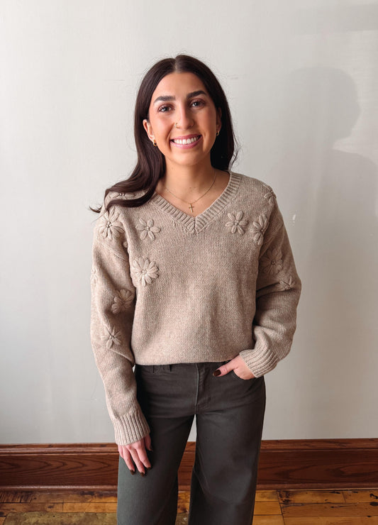 A woman wearing a beige knit sweater with raised floral patterns on the chest and sleeves, paired with dark gray pants, standing against a plain off-white wall.