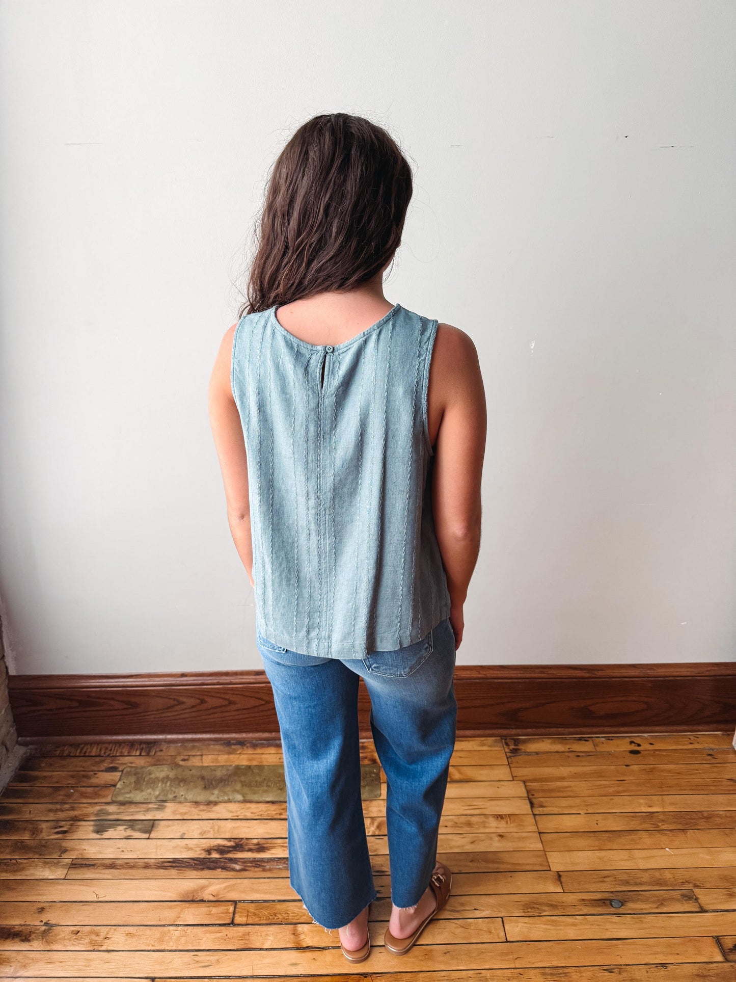 Person wearing a light blue sleeveless top and jeans standing against a white wall.