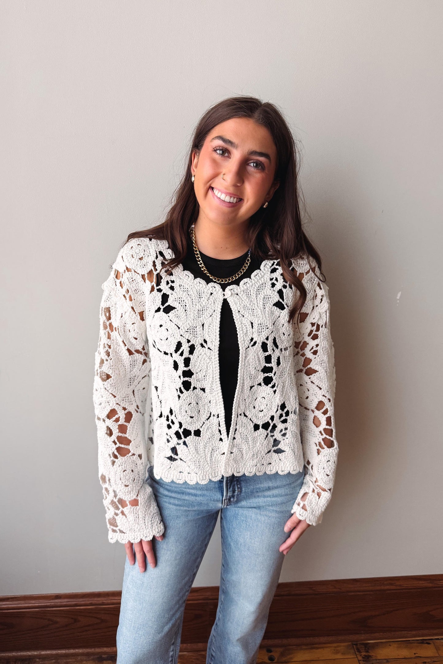 Woman wearing a white lace top and blue jeans against a plain wall.