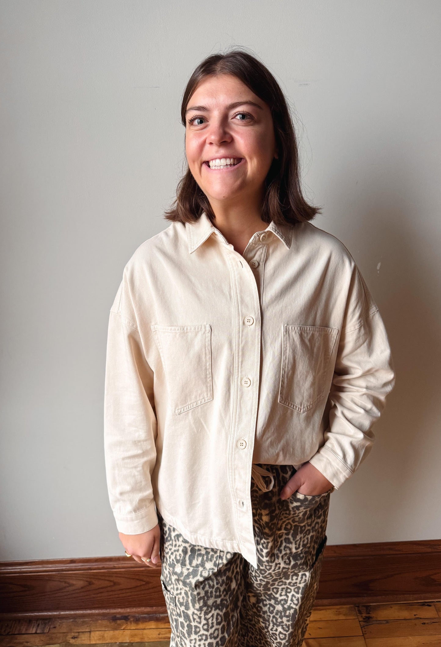 A woman smiling and looking up, wearing a light beige long-sleeve button-up shirt and leopard-print pants, standing against a plain light gray wall with a wooden baseboard and floor.