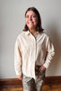 A woman smiling and looking up, wearing a light beige long-sleeve button-up shirt and leopard-print pants, standing against a plain light gray wall with a wooden baseboard and floor.