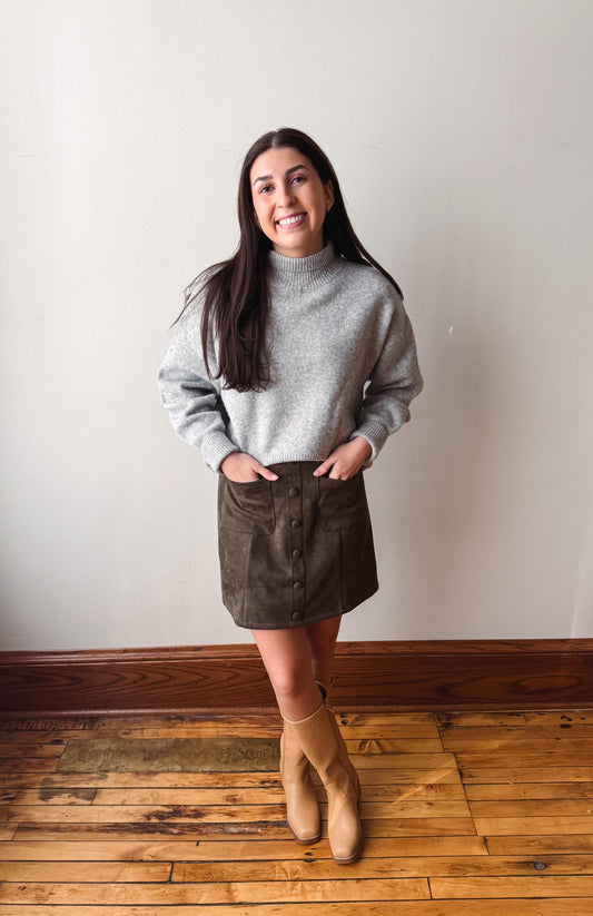 A woman modeling a light gray mock neck knit sweater and a dark brown corduroy mini skirt, paired with tan ankle boots, standing on a wooden floor against a plain off-white wall.