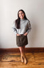 A woman modeling a light gray mock neck knit sweater and a dark brown corduroy mini skirt, paired with tan ankle boots, standing on a wooden floor against a plain off-white wall.