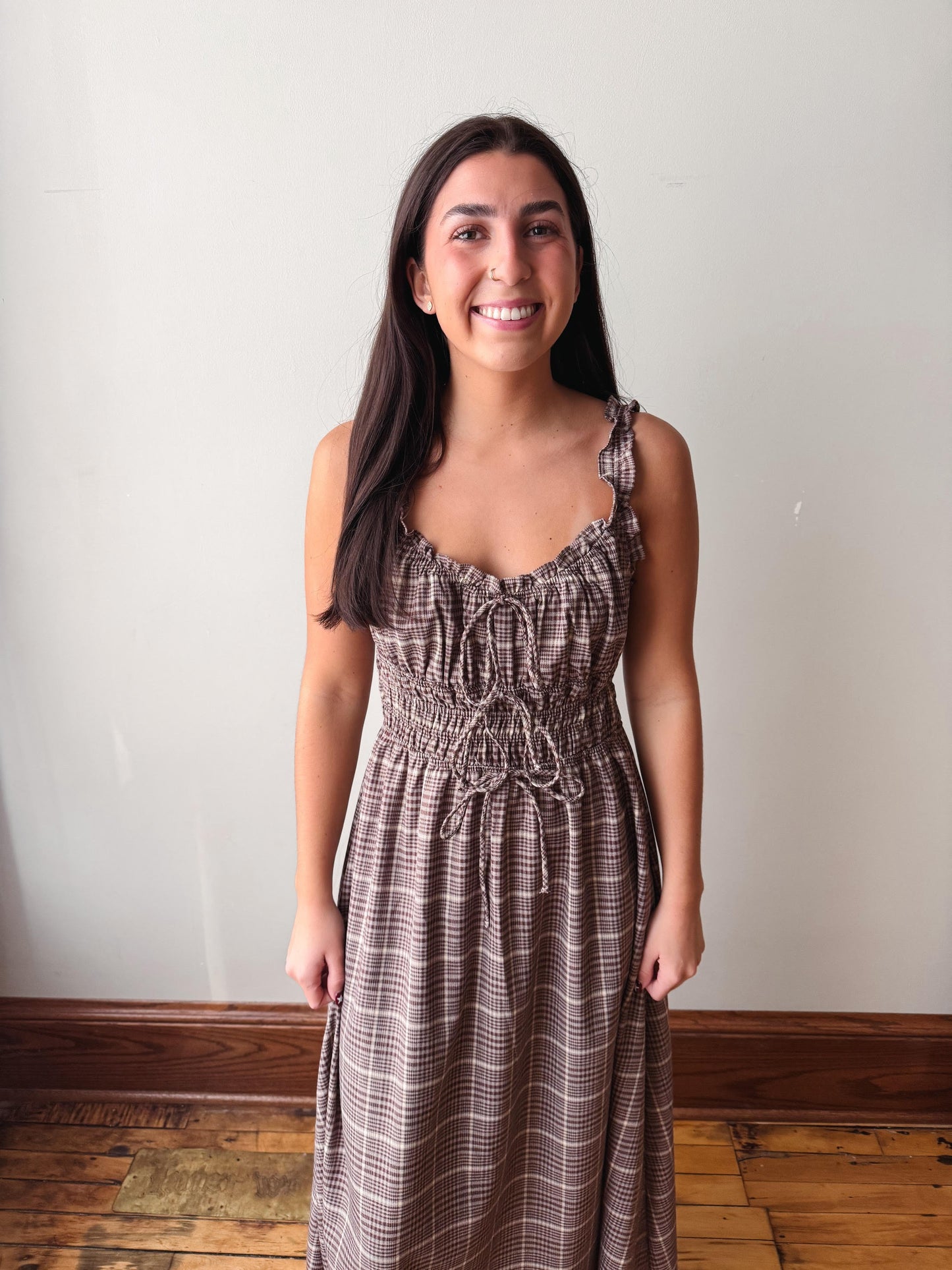A smiling young woman with long dark hair stands indoors wearing a sleeveless brown and beige plaid midi dress with a smocked bodice and thin shoulder straps, set against a plain off-white wall and wooden floor.