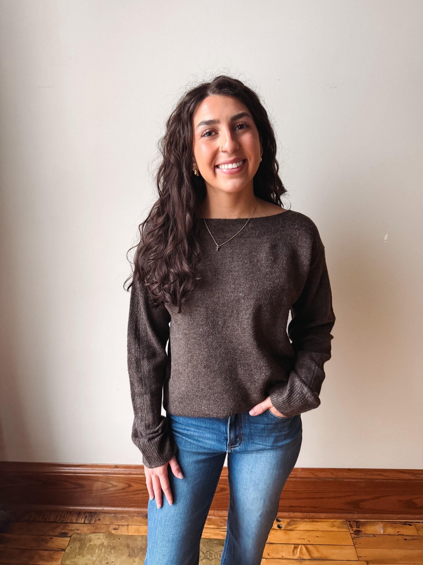 A woman with long, wavy dark brown hair stands smiling in front of a plain beige wall, wearing a dark brown knit sweater and blue jeans.