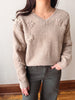 A woman wearing a beige ribbed knit sweater with raised daisy-like floral patterns on the front and sleeves, paired with dark gray pants, standing indoors against a light wall and wood floor.