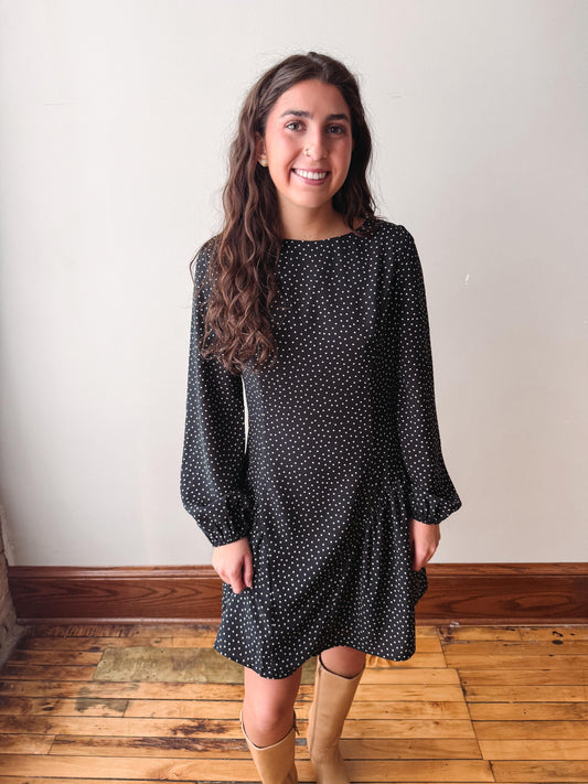 A woman wearing a black long-sleeve dress with a small white polka dot pattern, standing in front of a plain off-white wall on a wooden floor.