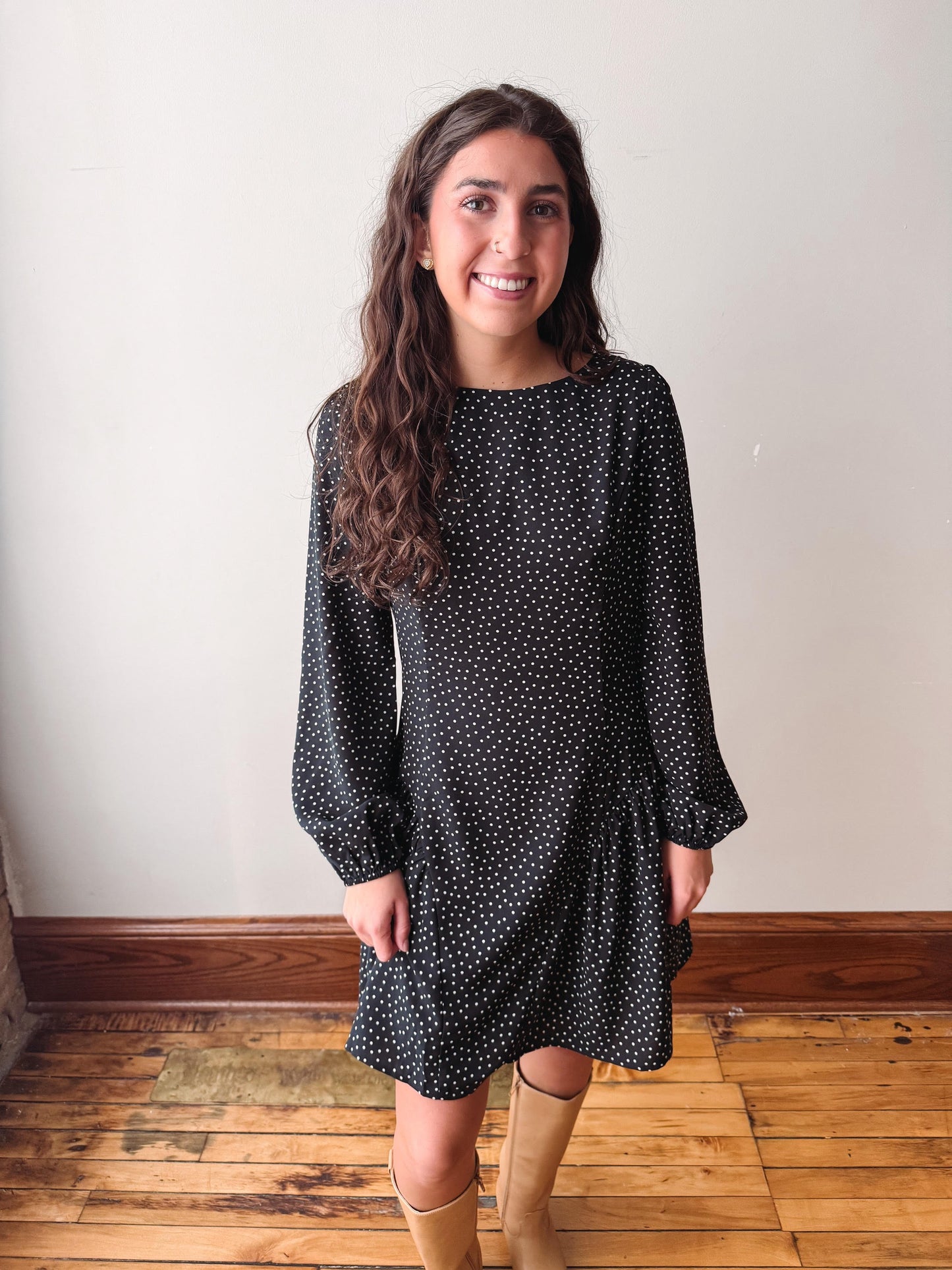 A woman wearing a black long-sleeve dress with a small white polka dot pattern, standing in front of a plain off-white wall on a wooden floor.