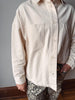A person wearing an oversized off-white linen button-up shirt with two chest pockets, paired with leopard-print pants, standing indoors against a gray wall and wooden floor.