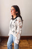 Woman wearing a white lace cardigan over a black top and blue jeans against a plain wall.
