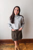 A woman modeling a light gray knit mock-neck sweater and a dark brown button-front skirt, standing on a wooden floor against a plain off-white wall.