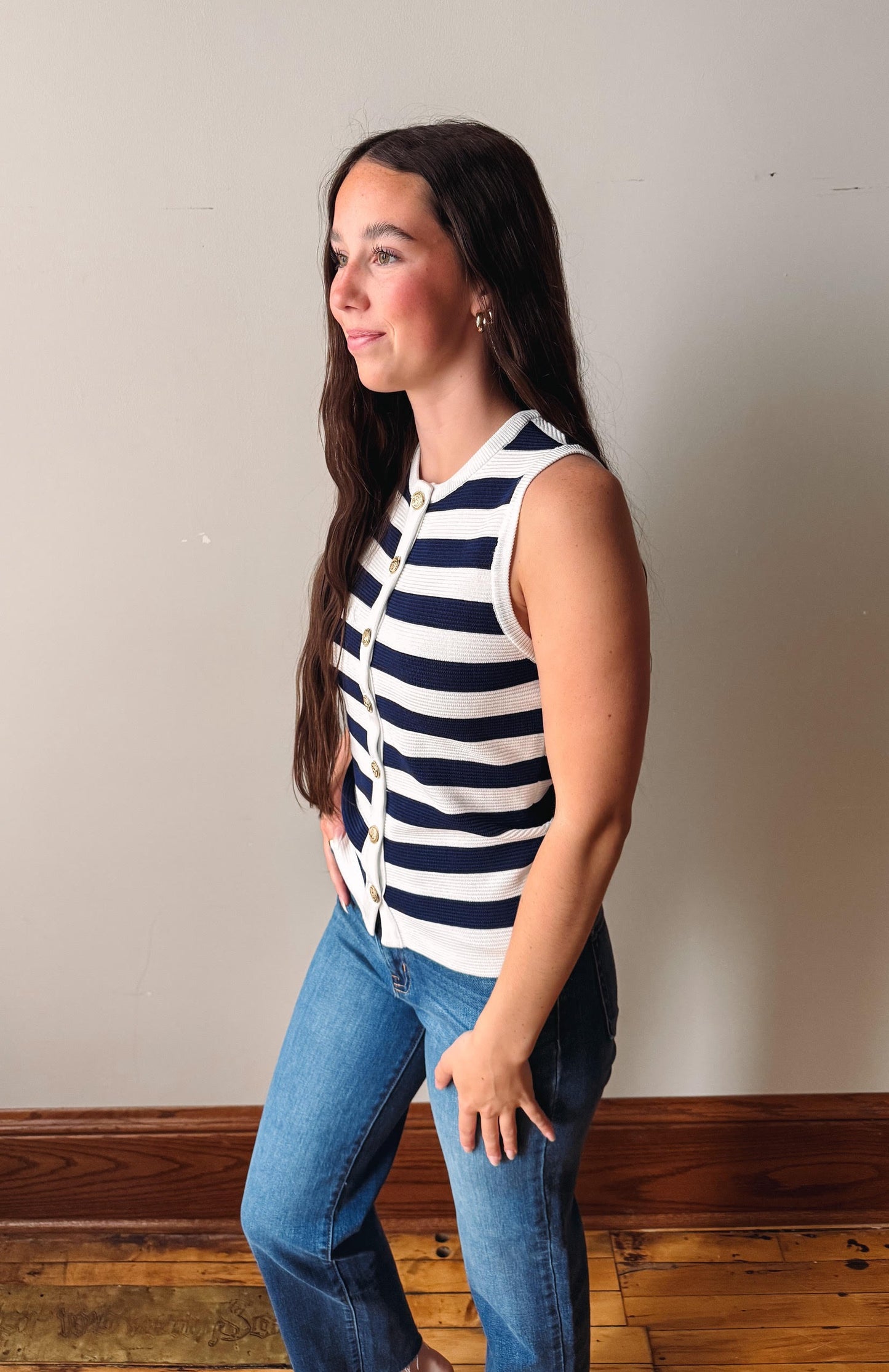 Woman wearing a navy and white striped sleeveless top and blue jeans against a plain wall.
